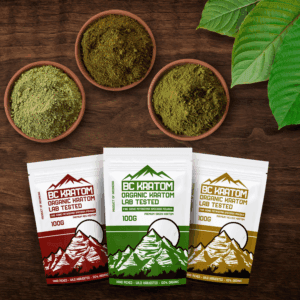 three 100g BC Kratom bags laying on a wooden table beside 3 different bowls of kratom powder